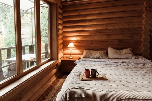 Rustikales Interieur eines Blockhaus-Schlafzimmers. Gemütliches Bett am großen Fenster. Frühstück auf einem Tablett im Hotel.